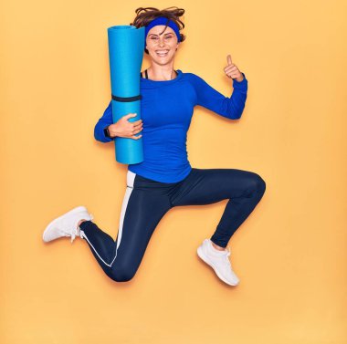 Genç ve güzel sporcu kadın mutlu gülümsüyor. Yüzünde tebessümle yoga minderini tutarak zıplıyorum. Tamam işareti yapıyorum. Başparmaklarımla izole edilmiş sarı arka plan üzerinde..