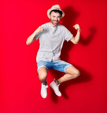 Genç, yakışıklı, günlük kıyafetler giyen ve şapkalı mutlu bir İspanyol. Yüzünde tebessümle zıplıyor, kırmızı arka planda yumruklar havada kutluyor..