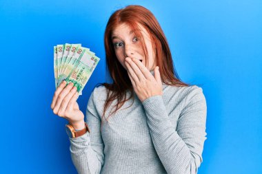 50 Hong Kong doları tutan genç, kızıl saçlı kız. Ağzı ağızla kaplayan banknotlar. Hata yapmaktan korkuyor. şaşırmış ifade 