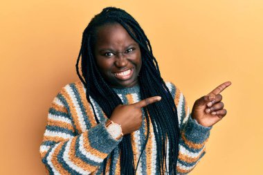 Young black woman with braids wearing casual winter sweater pointing aside worried and nervous with both hands, concerned and surprised expression 