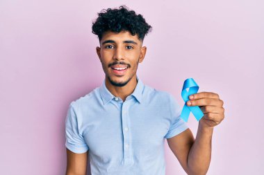 Genç Arap yakışıklı adam mavi kurdele tutuyor. Pozitif ve mutlu görünüyor. Duruyor ve gülümsüyor. 