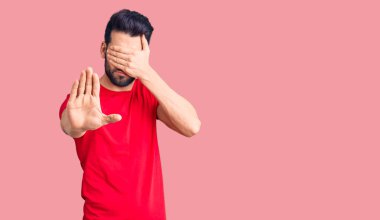 Sakallı, sıradan tişörtler giyen, elleriyle gözlerini kapatan, üzgün ve korku dolu hareketler yapan genç yakışıklı bir adam. utangaç ve olumsuz kavram. 