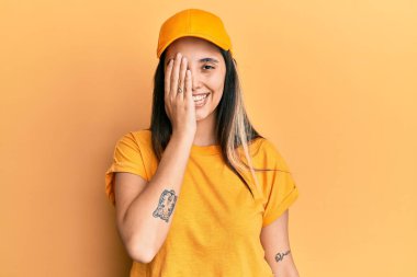 Teslimat üniforması giyen ve şapkalı genç İspanyol kadın bir gözünü eliyle kapatıyor, yüzünde kendine güvenen bir gülümseme ve sürpriz bir duygu var.. 