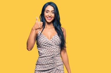 Yazlık elbise giyen güzel İspanyol kadın mutlu ve pozitif gülümsüyor, başparmağıyla mükemmel ve onay işareti yapıyor. 