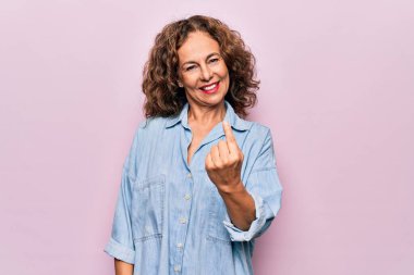 Orta yaşlı, güzel bir kadın, sıradan bir kot gömlek giyiyor pembe arka planda duruyor.