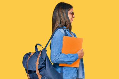 Sırt çantası takan ve elinde kitap tutan genç Latin bir kız. Profil profili için doğal bir yüz ve kendine güvenen bir gülümsemeyle poz veriyor.. 