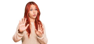 Kış süveteri giyen genç, kızıl saçlı bir kadın. Avuçlarını hareket ettiriyor. Korkmuş ve iğrenç bir ifadeyle reddediyor. Dur ve yasakla. 