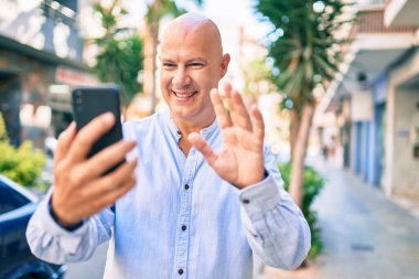 Orta yaşlı kel bir adam şehirde akıllı telefon kullanarak video araması yaparken mutlu gülümsüyor..