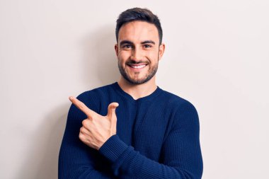 Sakallı, sıradan bir süveter giyen, beyaz arka planda durup gülümseyen, eli ve parmağı yanda olan genç yakışıklı bir adam.