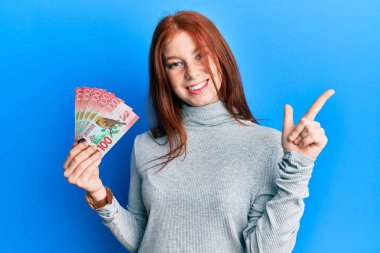 Genç kızıl saçlı kız elinde 100 yeni Zelanda doları banknotuyla gülümsüyor mutlu bir şekilde elini ve parmağını kenarı işaret ediyor. 