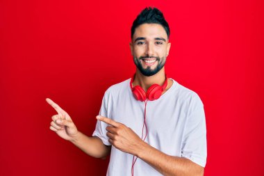 Young man with beard listening to music using headphones smiling and looking at the camera pointing with two hands and fingers to the side. 