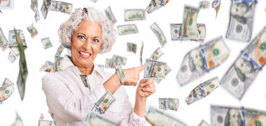 Yaşlı, gri saçlı, günlük kıyafetler giyen, gülümseyen ve kameraya bakan iki eliyle ve parmaklarıyla yüzünü gösteren bir kadın..