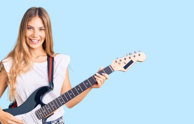 Güzel, beyaz, sarı saçlı, elektro gitar çalan güzel bir kadın. Pozitif ve mutlu görünüyor. Kendine güvenen bir gülümsemeyle dişlerini gösteriyor. 