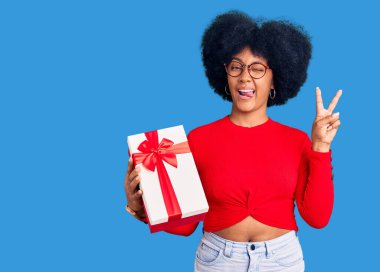 Genç Afrikalı Amerikalı kız elinde hediyeyle kameraya gülümseyen ve parmaklarıyla zafer işareti yapan. İki numara.. 