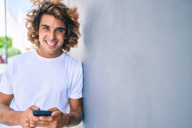 Duvara dayanmış akıllı telefon kullanan, mutlu gülümseyen genç İspanyol adam.