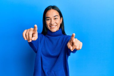 Kış süveteri giymiş genç Asyalı bir kadın seni ve kamerayı işaret ediyor, pozitif ve neşeli gülümsüyor. 