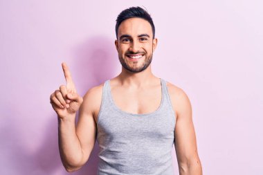 Sakallı, pembe arka planda kolsuz tişört giyen genç yakışıklı bir adam bir fikir veya soruyla gülümsüyor ve mutlu bir yüzle parmağını kaldırıyor.