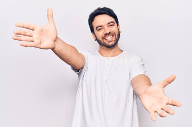 Gündelik giysiler giymiş genç İspanyol adam kameraya bakıp sarılmak için kollarını açmış gülümsüyor. Mutluluğu kucaklayan neşeli bir ifade.. 