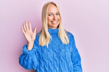 Kışın yün kazak giyen genç sarışın kız gülümseyerek kendinden emin ve mutlu gülümserken parmaklarıyla beş numarayı gösteriyor.. 