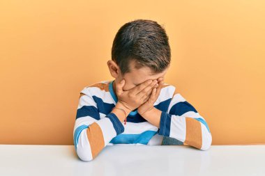 Kafkasyalı sevimli bir çocuk, gündelik giysiler giyip masaya oturmuş, suratını ellerle kapatmış, ağlarken. depresyon kavramı. 