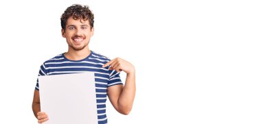 Genç, kıvırcık saçlı, boş afiş tutan, mutlu ve gururlu, kendini gösteren yakışıklı bir adam. 