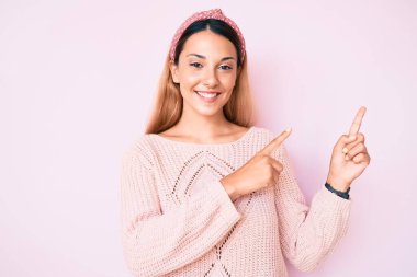 Genç esmer kadın pembe arka planda günlük kıyafetler giyiyor gülümsüyor ve kameraya bakıyor iki eliyle ve parmaklarıyla kenarı işaret ediyor.. 
