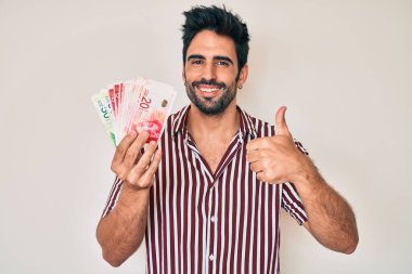 Sakallı yakışıklı İspanyol adam İsrail şekelleri tutuyor mutlu ve pozitif gülümsüyor, başparmağıyla mükemmel ve onay işareti yapıyor. 