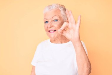 Son sınıf güzeli, mavi gözlü, gri saçlı, sarı arka planda klasik beyaz tişört giyen, gülümseyen, el ve parmaklarıyla 