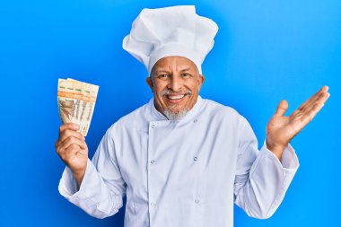Orta yaşlı, gri saçlı, profesyonel aşçı üniforması giyen, Norveçli krona banknotları tutan mutlu bir gülümsemeyle başarıyı kutlayan ve kazanan ifadeyle elini kaldıran bir adam. 