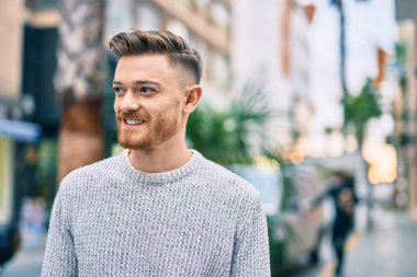 Genç beyaz adam gülümseyerek şehirde mutlu mesut duruyor..