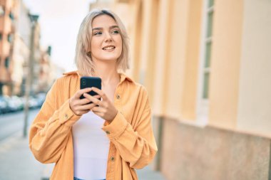 Genç sarışın kız şehirde akıllı telefon kullanarak mutlu bir şekilde gülümsüyor..