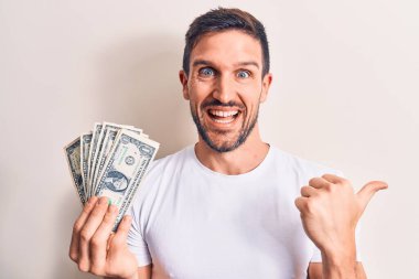 Genç yakışıklı bir adam, elinde bir deste banknotla soyutlanmış beyaz arka plan üzerinde başparmağını yukarı kaldırarak mutlu bir şekilde ağzı açık gülümsüyor.