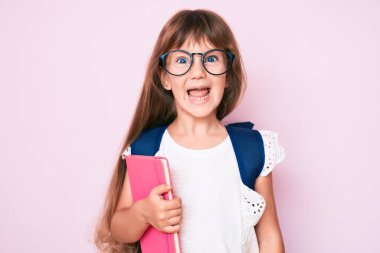 Uzun saçlı, öğrenci çantası takan, elinde kitap tutan, pozitif ve mutlu görünen, kendine güvenen gülümseyen, dişlerini gösteren beyaz bir kız çocuğu. 