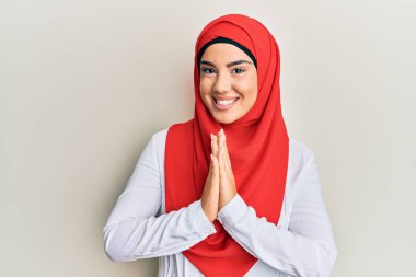 Young beautiful hispanic girl wearing traditional islamic hijab scarf praying with hands together asking for forgiveness smiling confident.  clipart