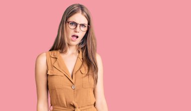 Şok olmuş yüzüne gözlük takmış genç, güzel, sarışın bir kadın şüpheci ve alaycı görünüyor. 