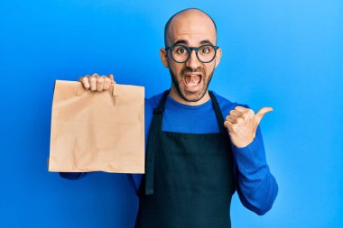 Garson üniforması giymiş genç İspanyol bir adam. Elinde paket poşeti var. Başparmağını kaldırıp gülümsüyor ve ağzı açık. 