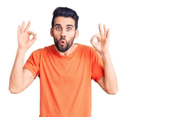 Sakallı, sıradan tişörtler giyen genç ve yakışıklı bir adam. Şaşırmış ve şaşırmış görünüyor. Parmaklarıyla onay sembolü yapıyor. çılgın ifade 