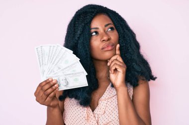 Dolarları tutan güzel Afrikalı bir kadın ciddi bir yüz ifadesiyle eli çenesinde soru soran, kafa karıştırıcı bir fikir hakkında düşünceli bir kadın. 