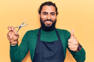 Berber önlüğü giymiş genç Arap adam mutlu ve pozitif gülümsüyor, başparmağıyla mükemmel ve onay işareti yapıyor. 