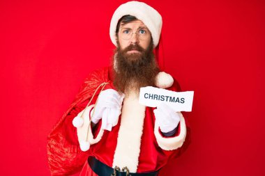 Uzun sakallı, Noel Baba kostümü giyen yakışıklı, genç, kızıl saçlı, yüzünde ciddi bir ifadeyle gevşemiş bir yılbaşı mesajı tutan adam. Basit ve doğal bir şekilde kameraya bakıyorsun.. 