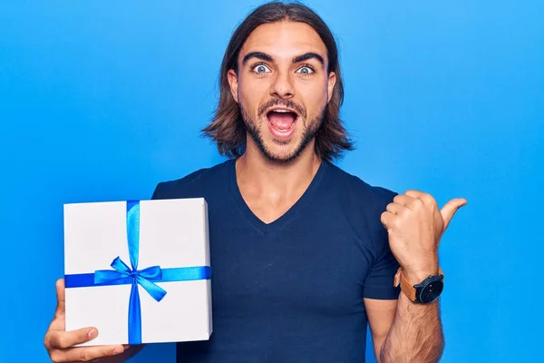 Jovem Homem Bonito Segurando Presente Apontando Polegar Até Lado Sorrindo — Fotografia de Stock