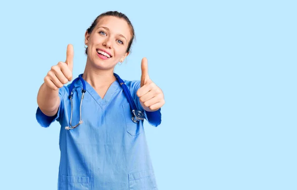 医師の制服を着た若い美しいブロンドの女性と手で積極的なジェスチャーを行うことを承認聴診器 親指アップ笑顔と成功のために幸せ 勝者のジェスチャー — ストック写真