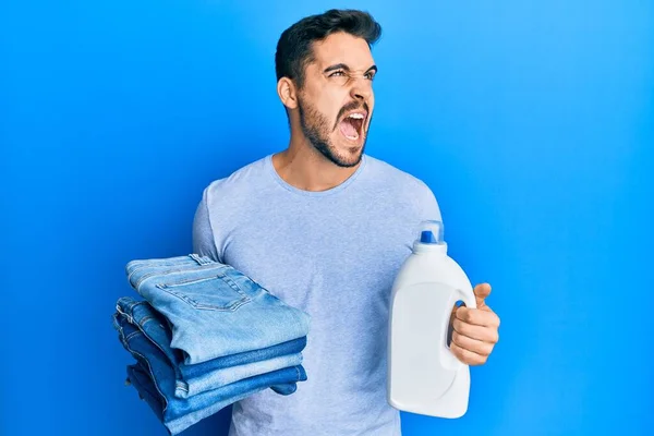 Joven Hombre Hispano Sosteniendo Pantalones Vaqueros Doblados Botella Detergente Enojado —  Fotos de Stock