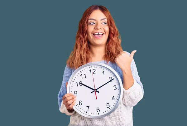 Young Latin Woman Holding Big Clock Pointing Thumb Side Smiling — Stock Photo, Image