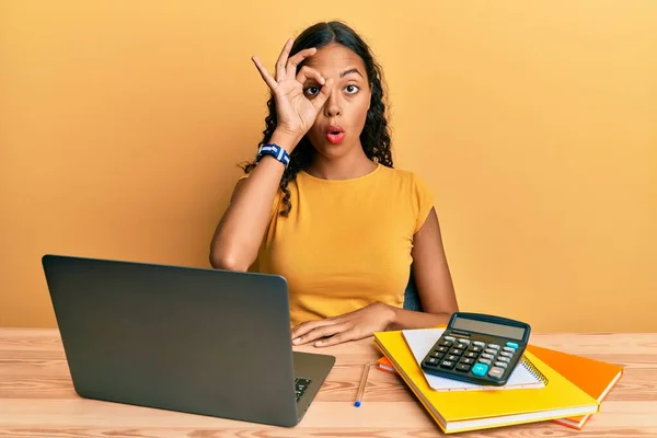 Young african american girl working at the office with laptop and calculator doing ok gesture shocked with surprised face, eye looking through fingers. unbelieving expression.