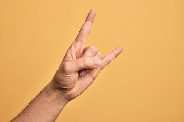 Hand of caucasian young man showing fingers over isolated yellow background gesturing rock and roll symbol, showing obscene horns gesture