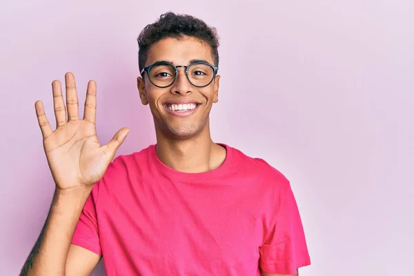 若いですハンサムなアフリカ系アメリカ人男性身に着けています眼鏡上のピンクの背景表示と指差す指番号5ながら笑顔自信と幸せ — ストック写真