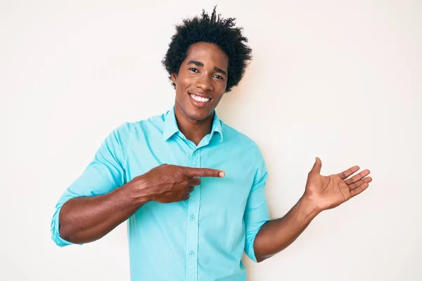 手で提示し 指で指している間 カジュアルな服を身に着けているアフロの髪を持つハンサムなアフリカ系アメリカ人男性は カメラに驚いて笑顔 — ストック写真