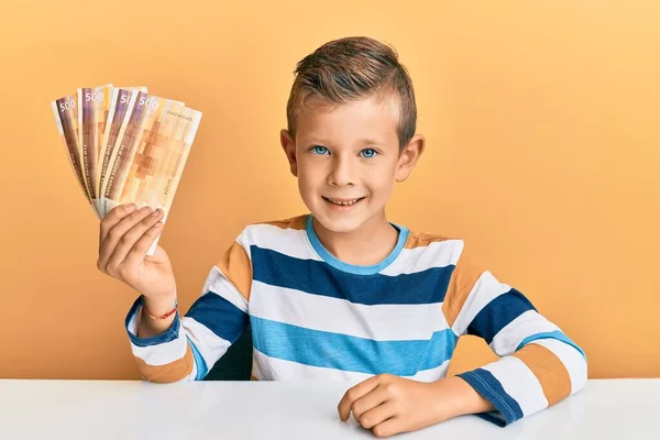 テーブルの上に座っている500ノルウェークローネの紙幣を保持している愛らしいCaucasian子供はポジティブで幸せな立ち姿で 自信を持って笑顔で歯を見せています — ストック写真