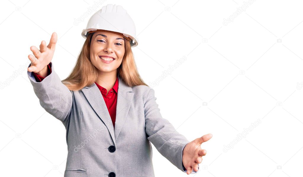 Beautiful young woman wearing architect hardhat looking at the camera smiling with open arms for hug. cheerful expression embracing happiness. 